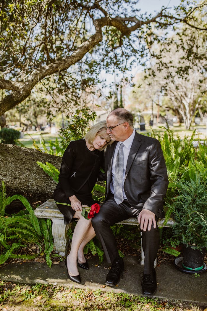 home-services-004 Elderly couple grieving with red roses, seated on an outdoor stone bench amidst greenery.