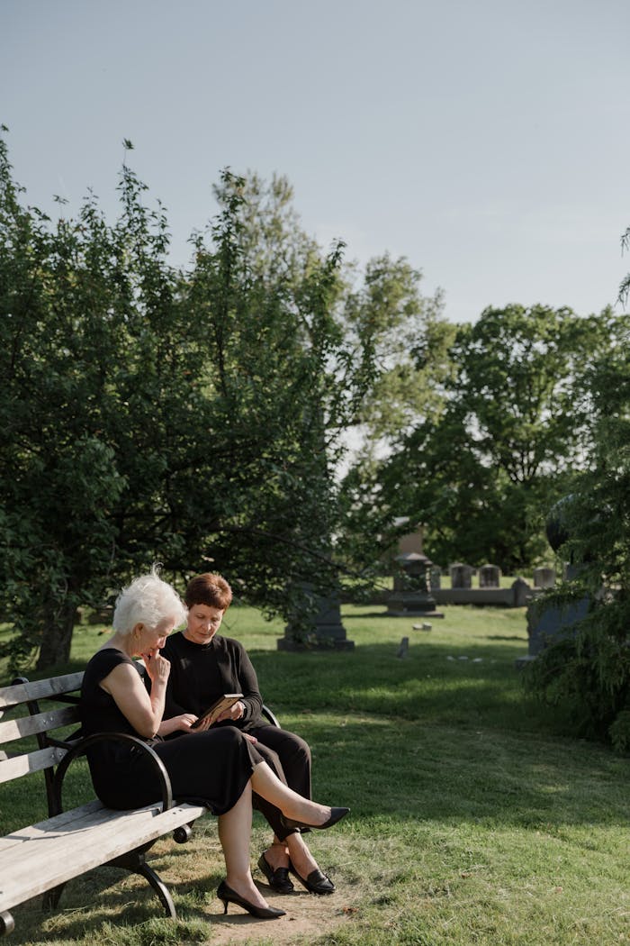 Two senior women sitting on a bench, mourning in a peaceful cemetery setting.