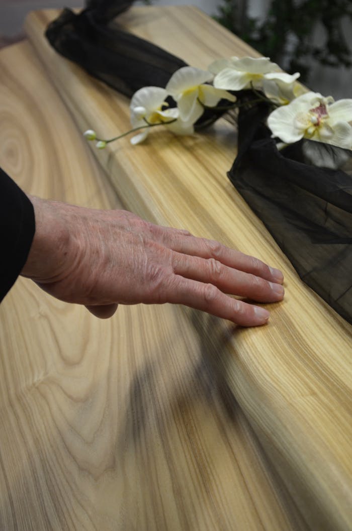 testimonials-img Close-up of a hand touching a wooden coffin adorned with flowers and black fabric, conveying a sense of mourning.